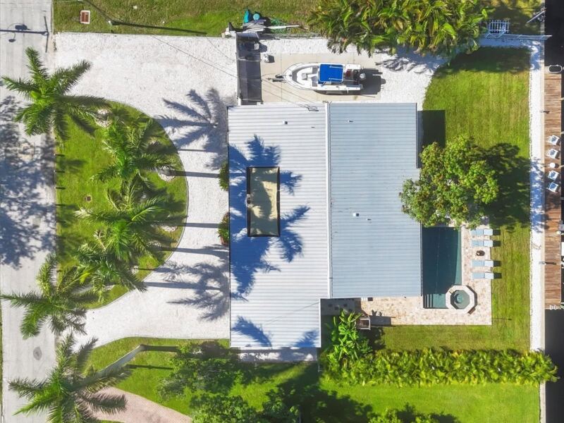 Aerial view of a canal-front home with a shell driveway, palms, lawn and dock
